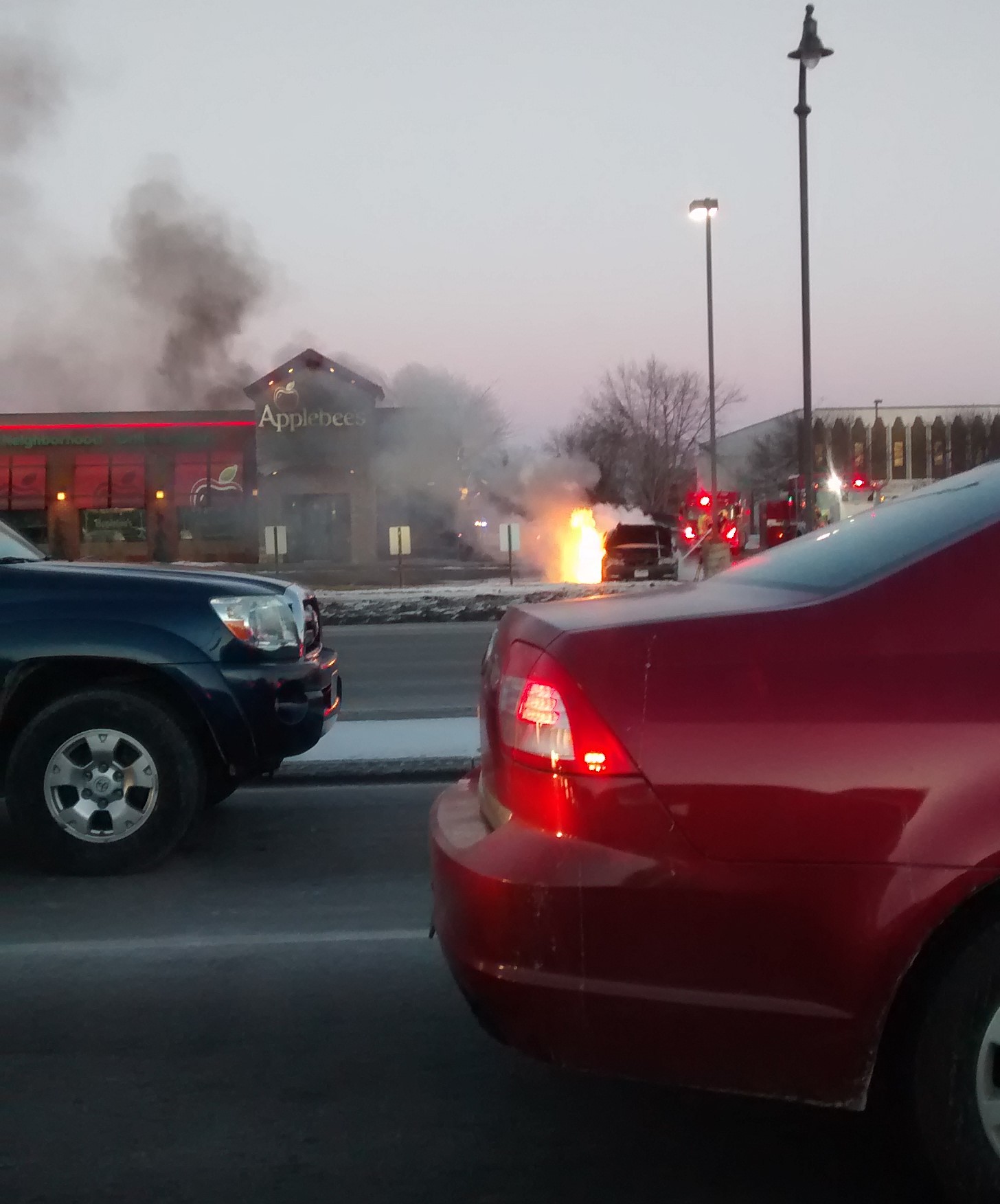 A car on fire in front of an Applebees.  Cars are patiently waiting in line to turn at a stoplight.  The scene has the feeling of the Wizard of Oz, Pay no attention to the man behind the curtain 